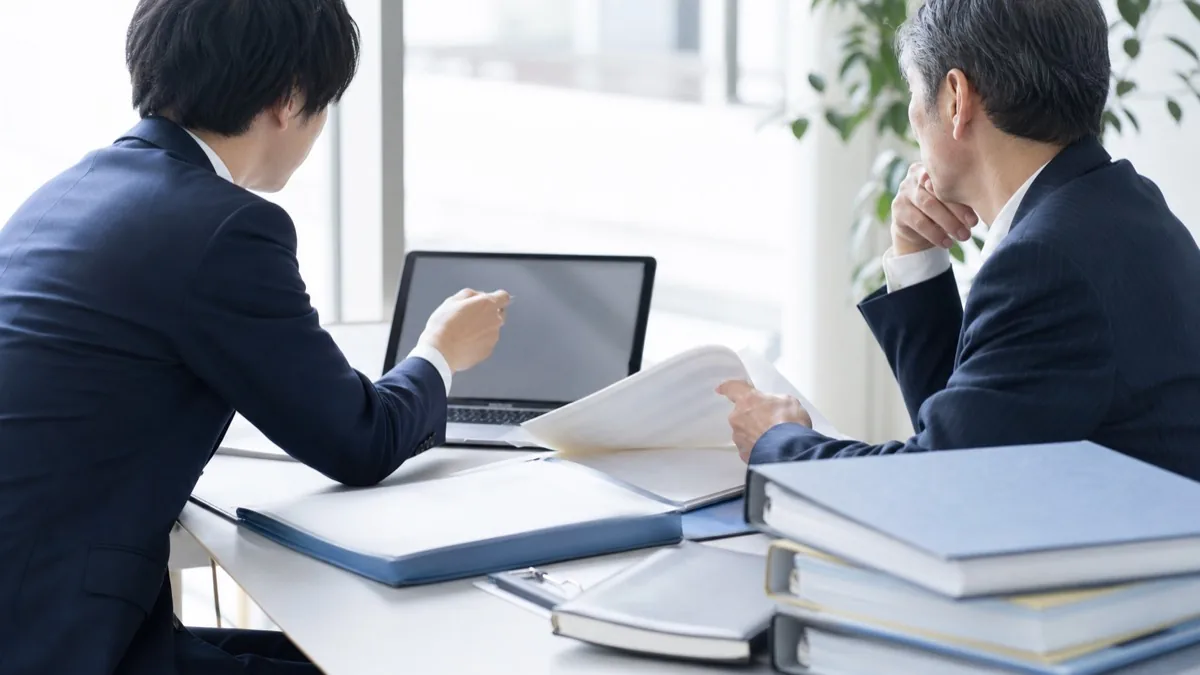 明るい会議室で、担当者と管理職がノートPCの画面と紙のファイル資料を見比べながら、社内チャットボットの導入を検討している様子